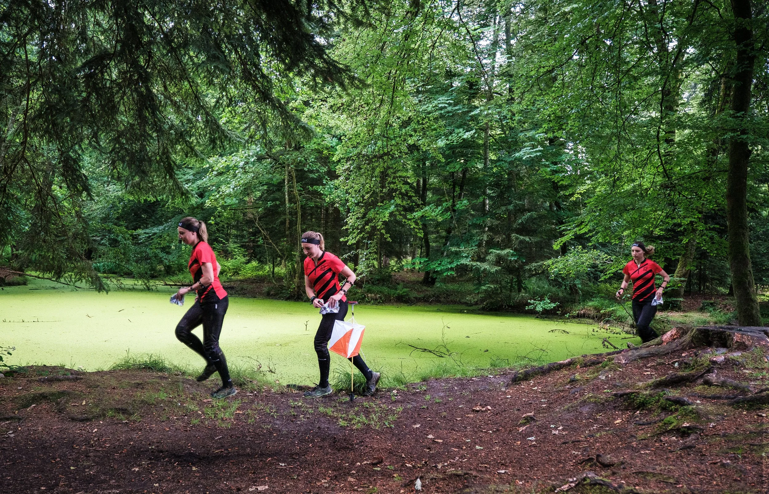 Drei Frauen beim Nachtwachen in einem grünen, bewaldeten Park. Sie tragen rote und schwarze Sportbekleidung und eine trägt einen Laufkegel.