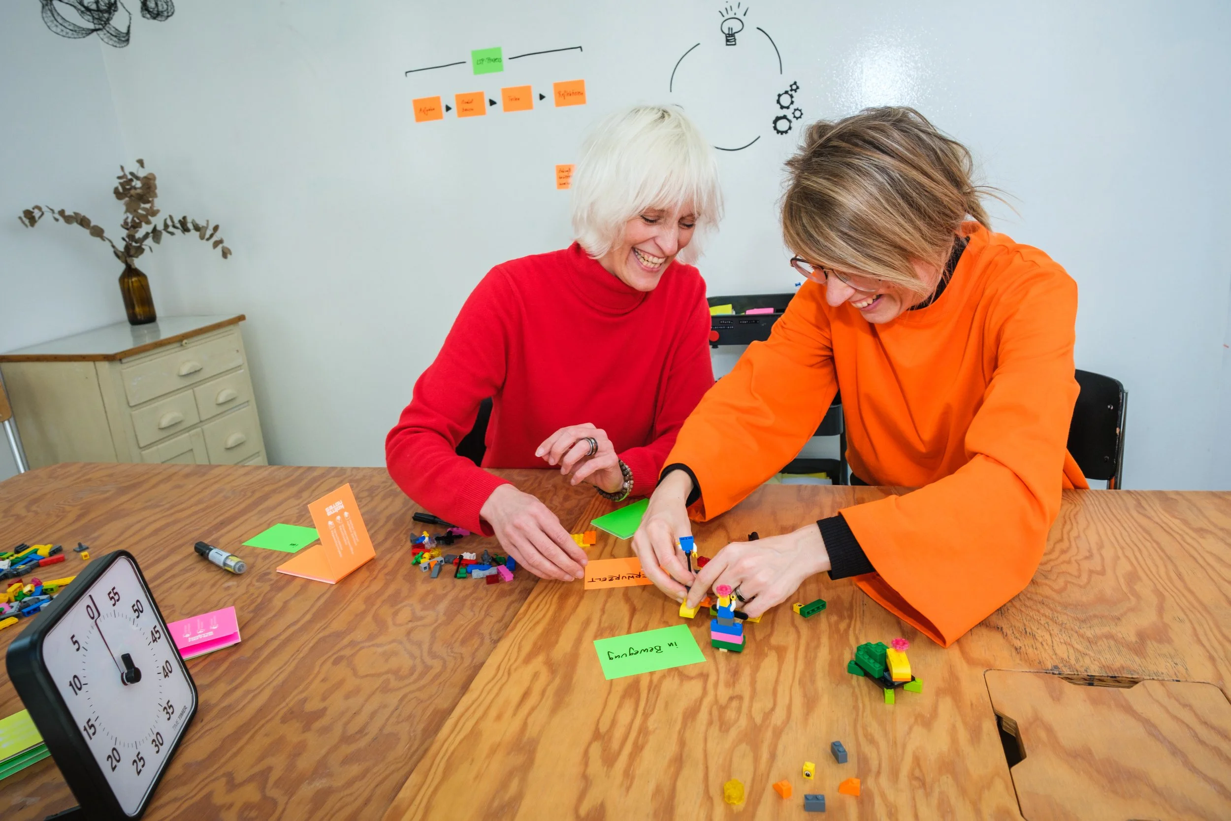 Zwei Frauen spielen mit LEGO-Steinen an einem Tisch, lachen und haben Spaß. Im Hintergrund eine weiße Tafel mit Post-its und Zeichnungen.