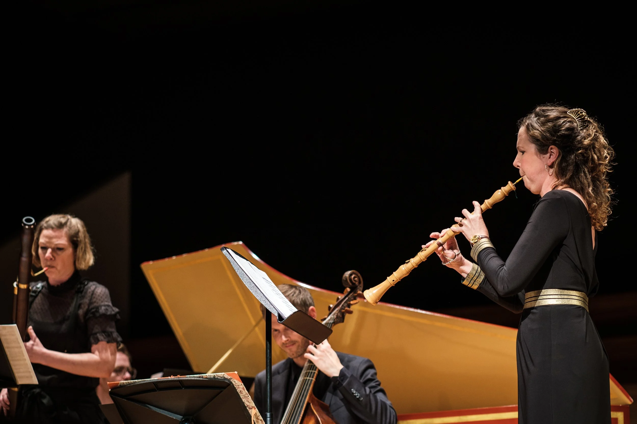 Musikerin spielt auf einer Flöte bei einem Orchesterkonzert, im Hintergrund sind andere Musiker und ein Klavier zu sehen.