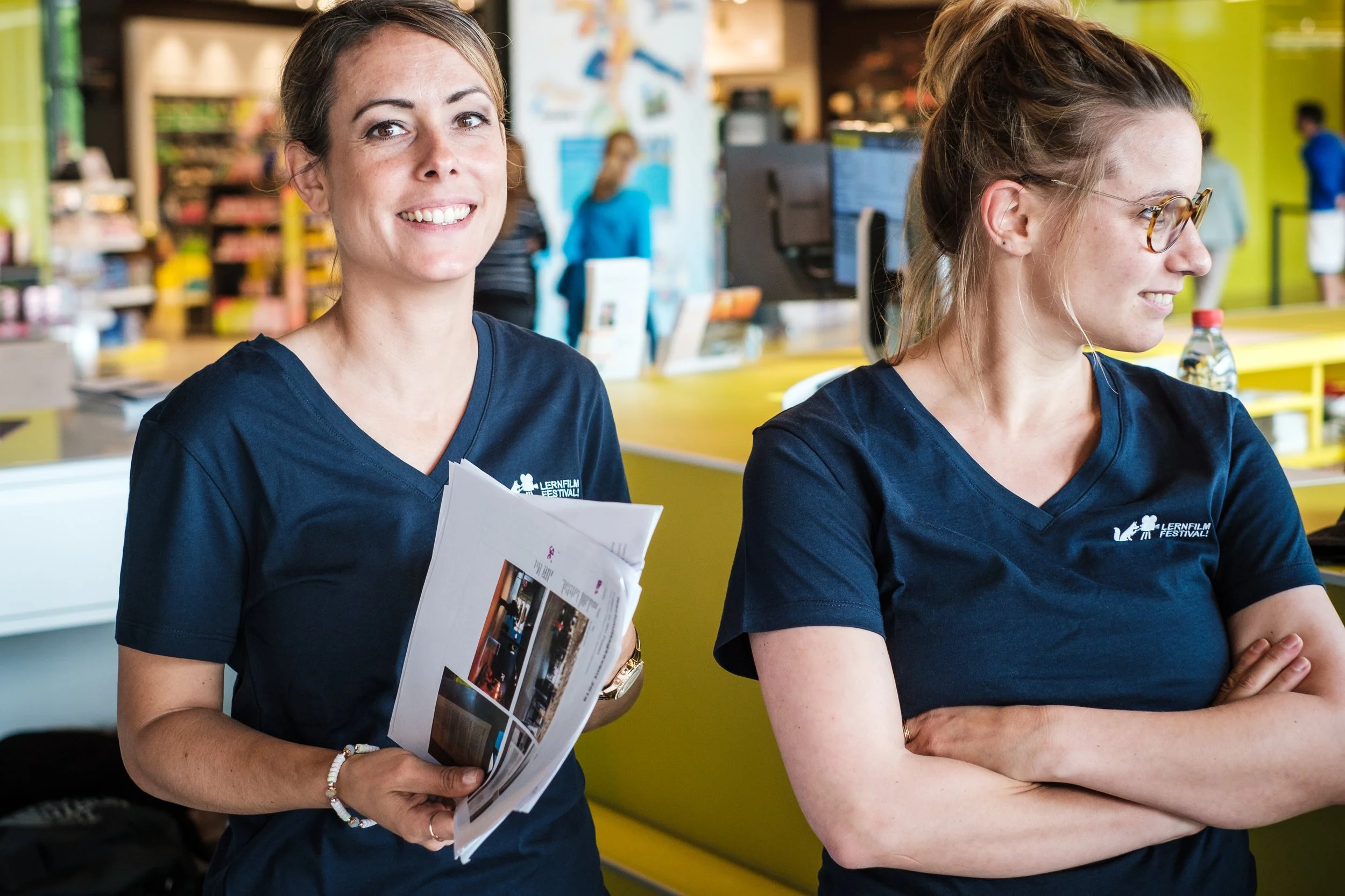 Zwei Frauen in blauen T-Shirts bei einem Bildungsevent, eine lächelnd und die andere mit verschränkten Armen, in einem gelben Raum mit Menschen im Hintergrund.