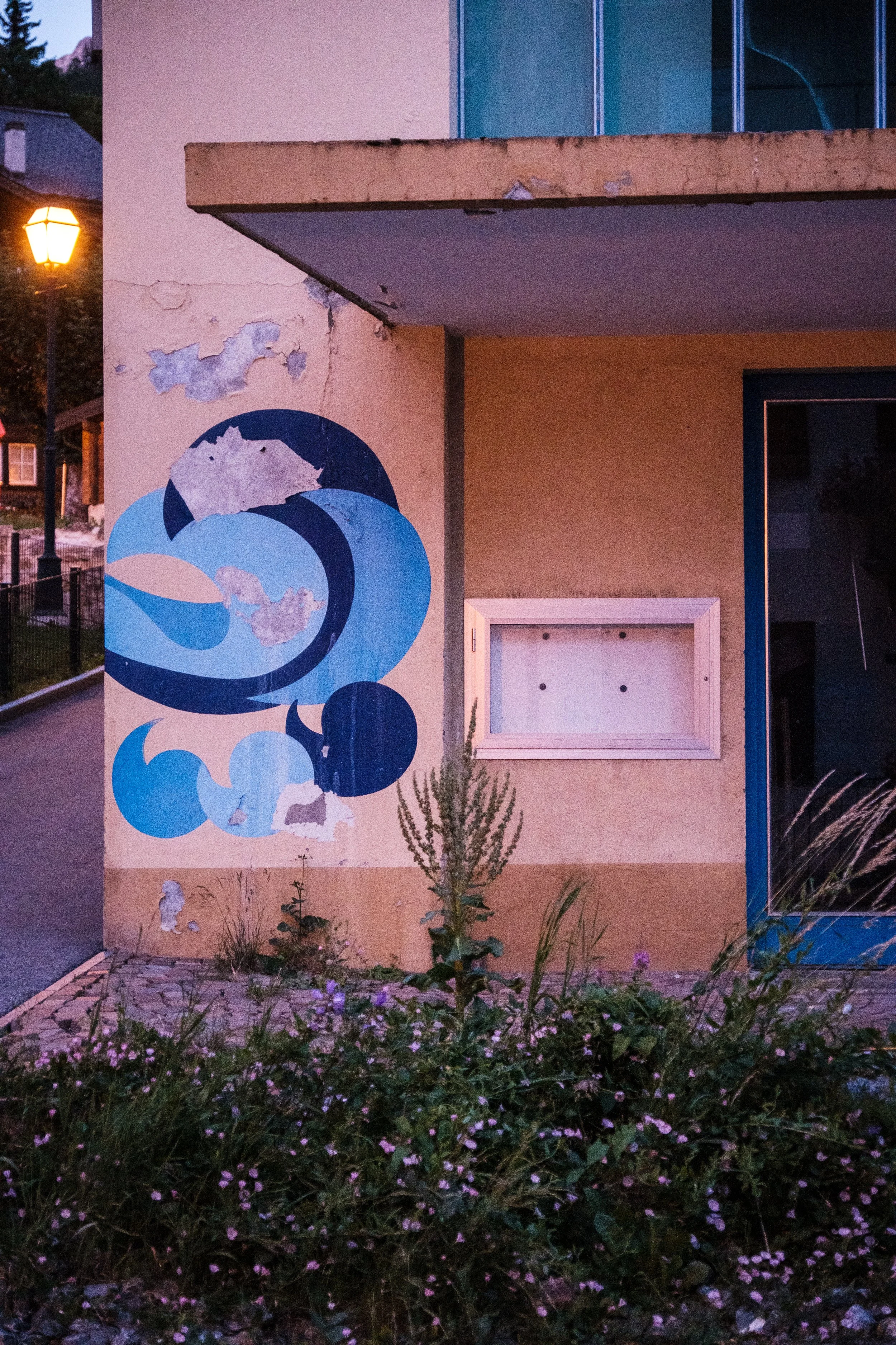Eine Wand mit einem blauen Wellenmuster und einigen beschädigten Stellen, umgeben von Pflanzen und Blumen im Vordergrund, mit zarten Lichter im Hintergrund.