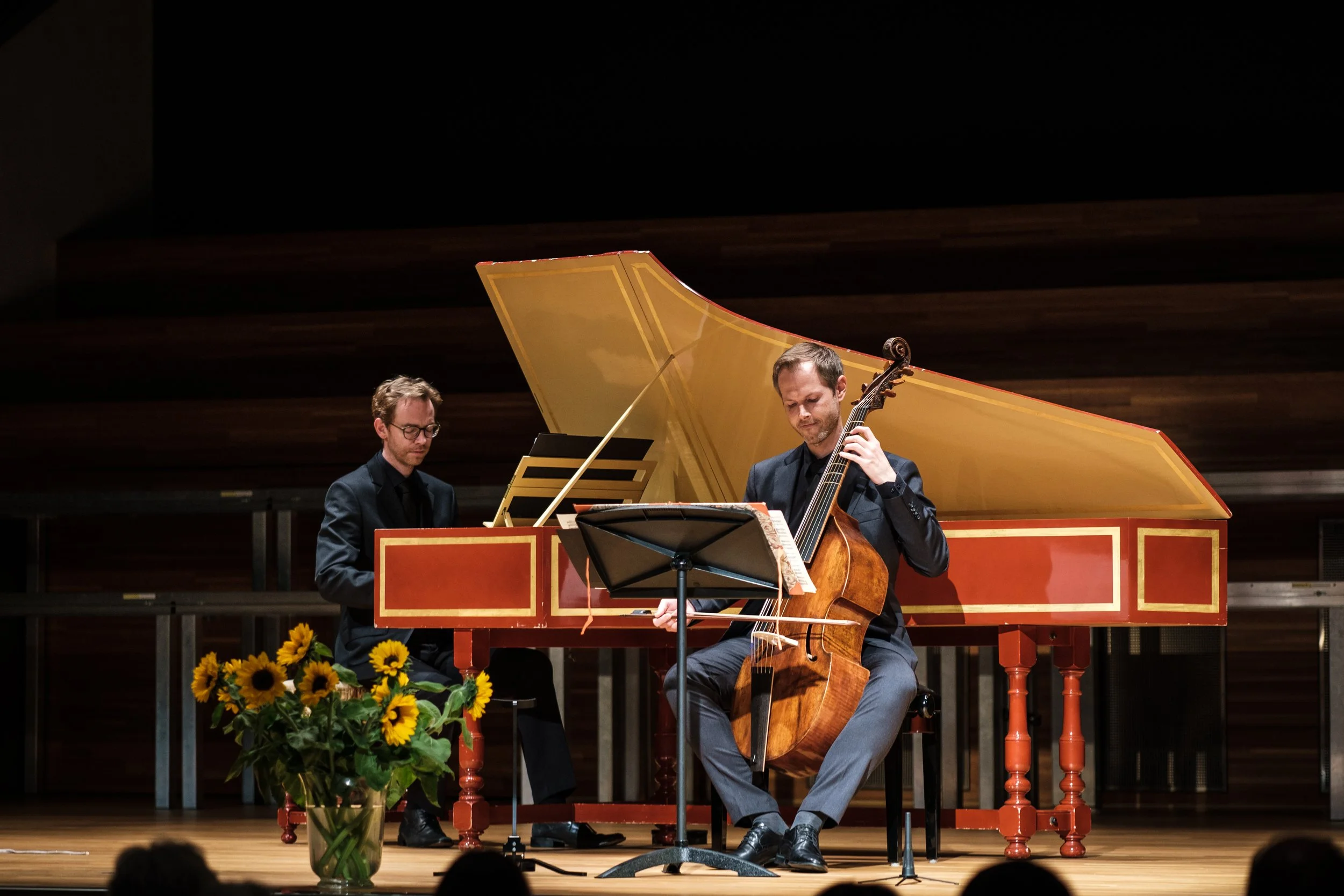 Zwei Musiker spielen auf einer Bühne: einer am Klavier und einer am Cello. Im Vordergrund steht ein Blumenstrauß mit Sonnenblumen.