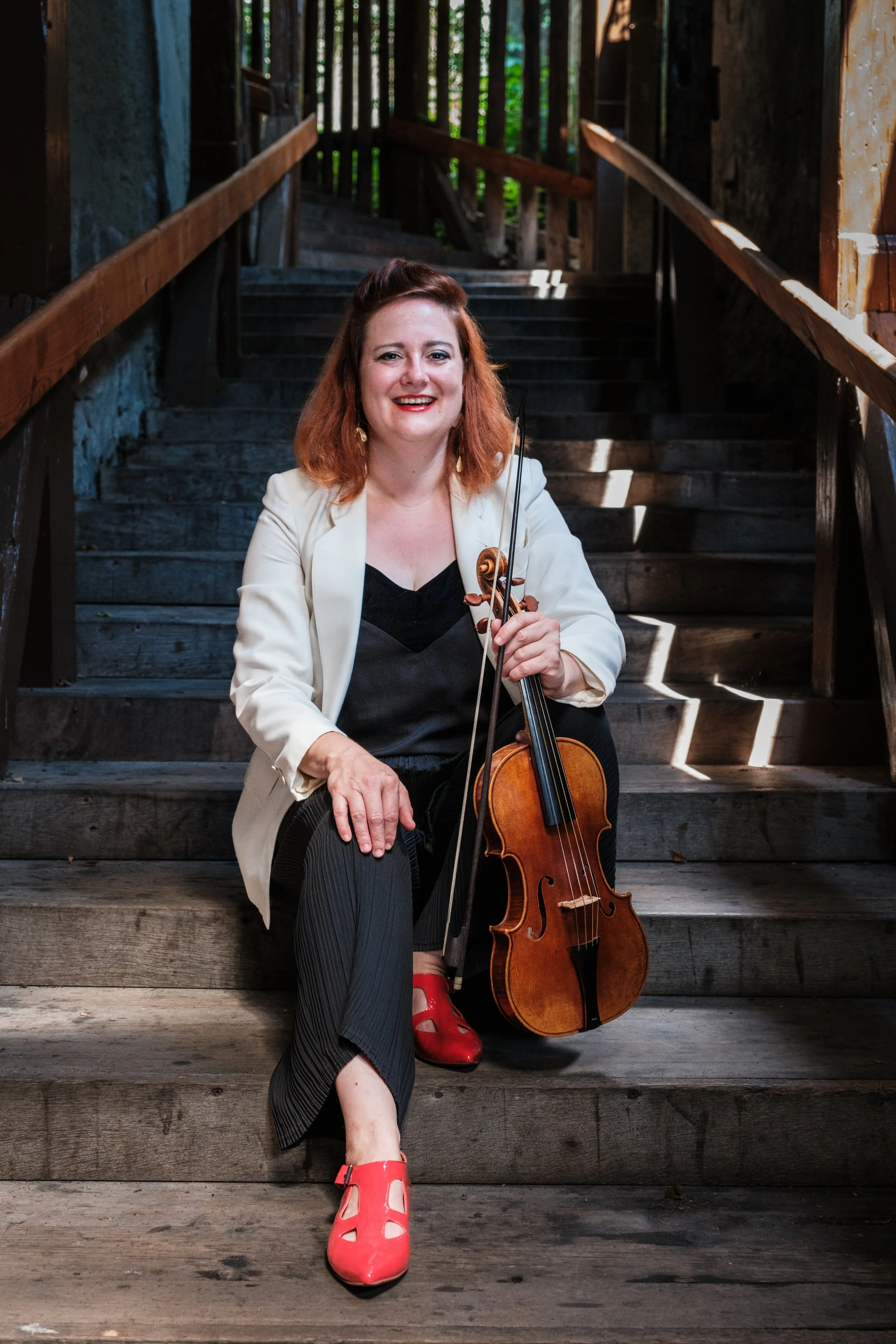 Eine Frau mit roten Haaren, in einem weißen Blazer und schwarzen Kleid, sitzt auf einer Holzstufe mit einem Violin in der Hand, im Hintergrund eine Holztreppe in einem dunklen Raum.