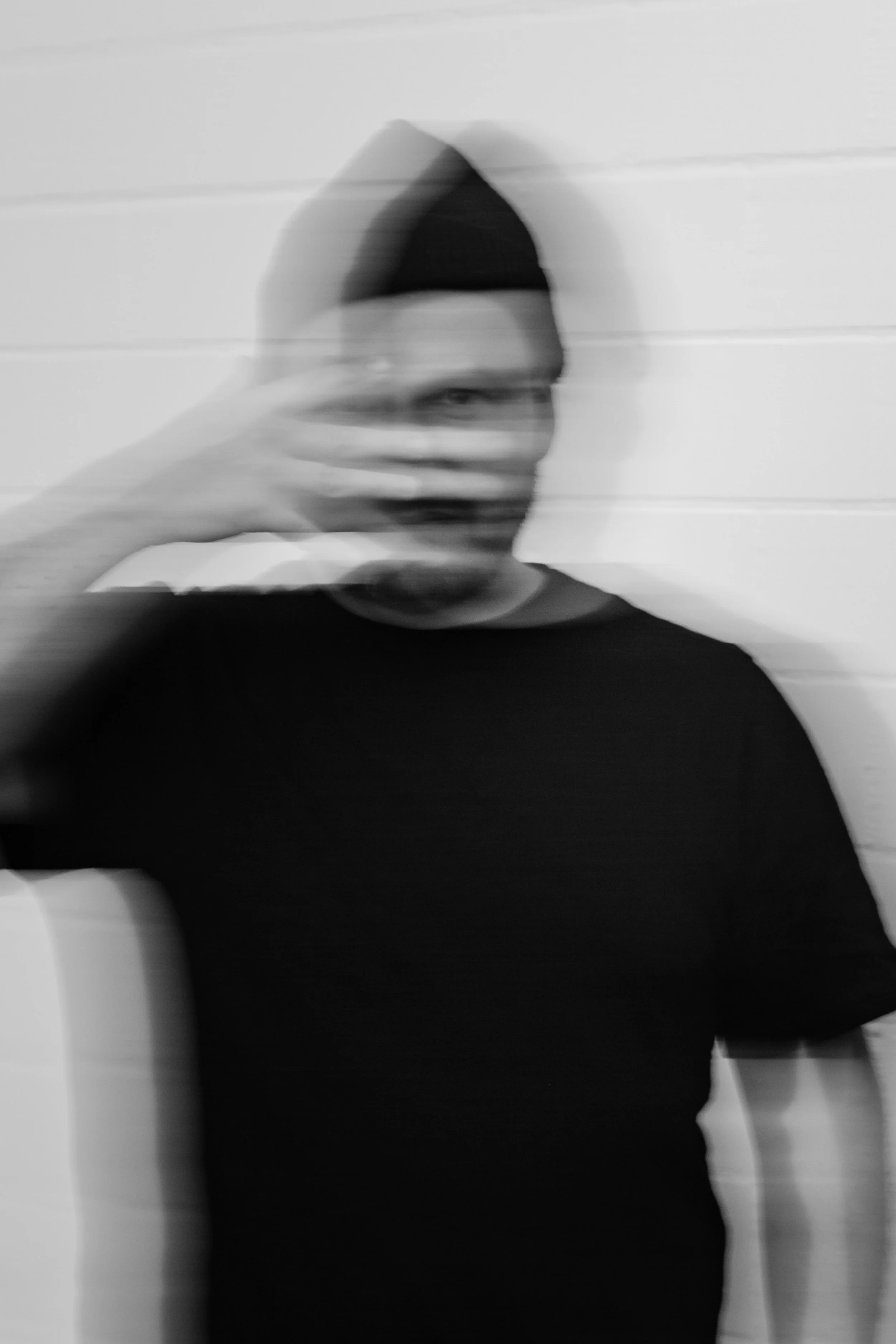 Schwarz-weiß-Fotografie eines Mannes in dunkler Kleidung, der eine schwarze Mütze trägt. Die Aufnahme ist absichtlich verschwommen, da der Mann sich bewegt oder die Kamera unscharf ist. Der Hintergrund besteht aus weißen Paneelen.