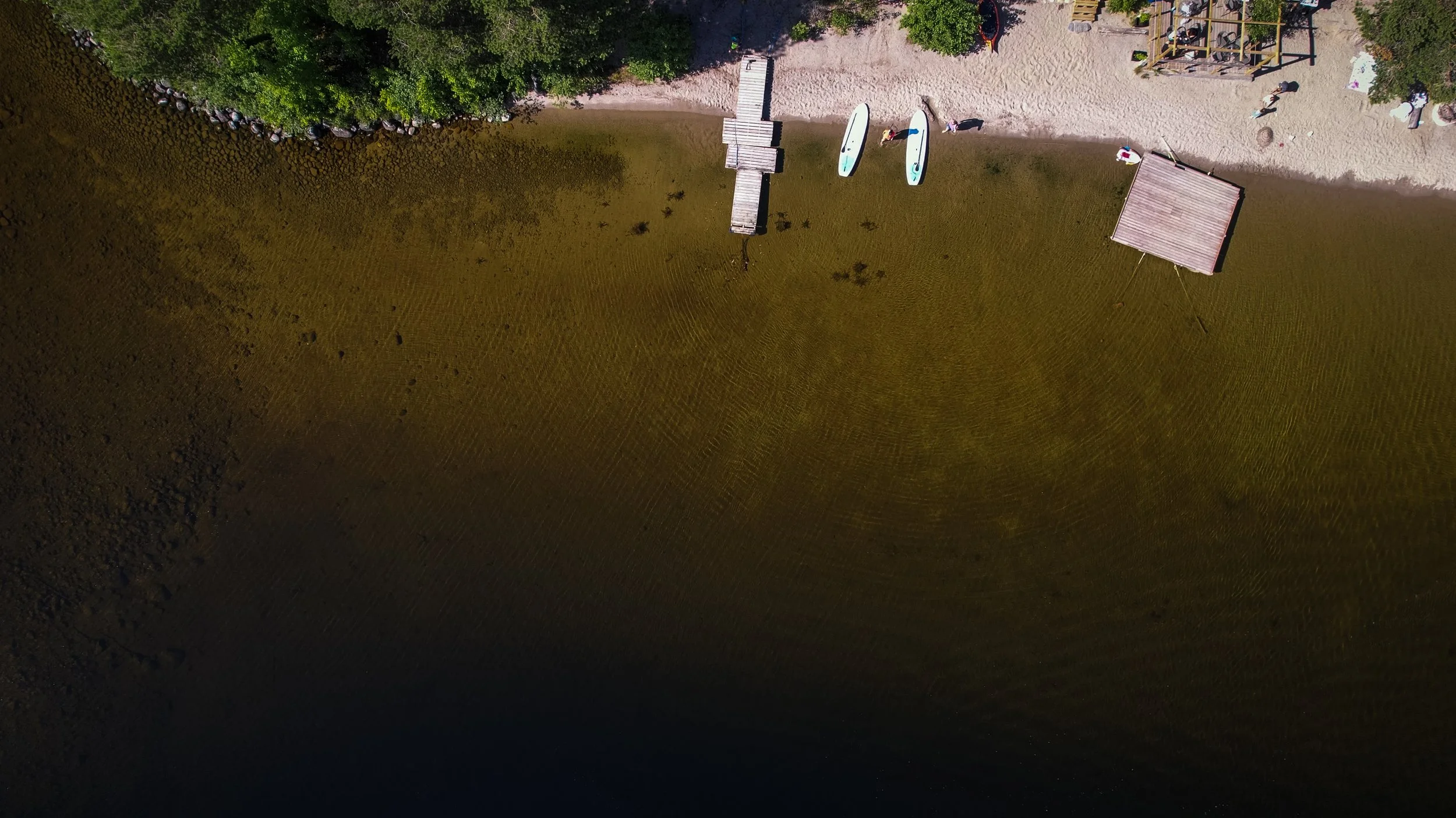 Vogelperspektive auf einen Strand mit zwei Surfbrettern, einer kleinen Holzbrücke, mehreren Menschen und einem Sandspielbereich.