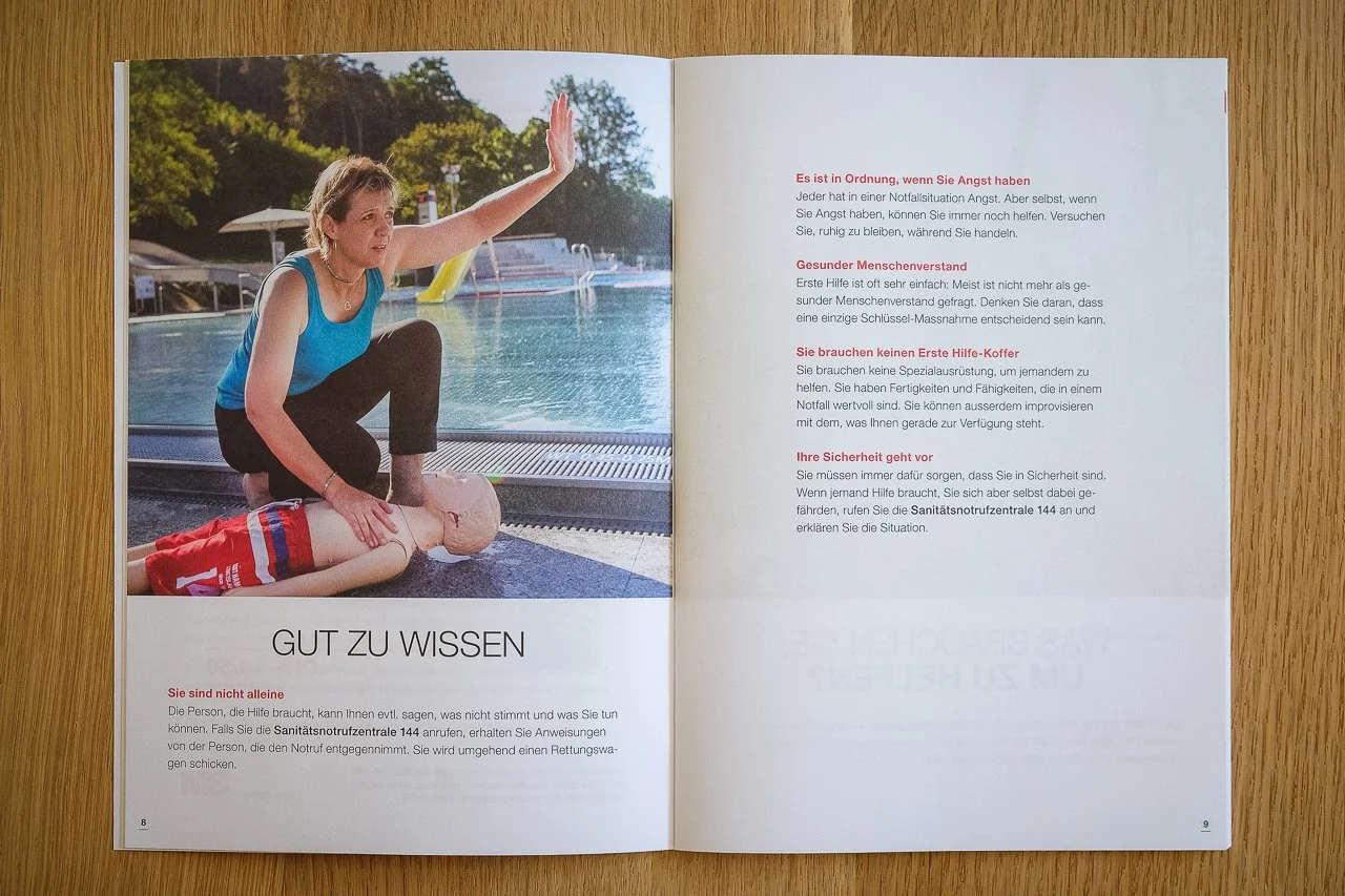 Offene Broschüre mit Bild eines Erwachsenen, der bei einem Pool eine Herz-Lungen-Wiederbelebung an einem Kind durchführt. Text auf der rechten Seite über Erste Hilfe und Sicherheit.