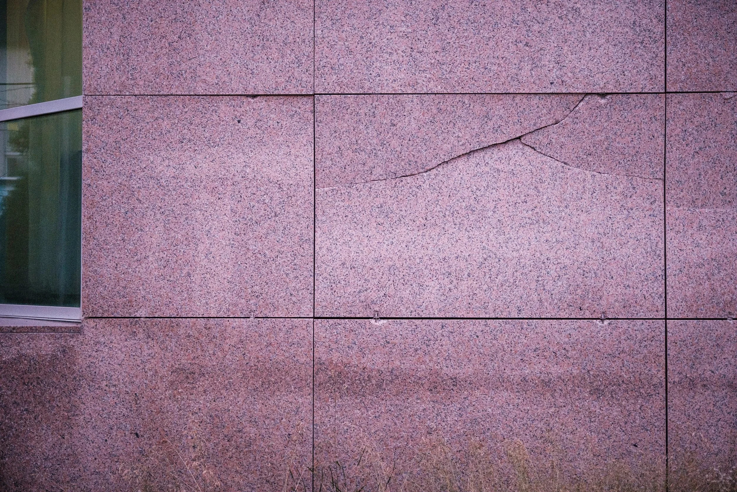 Pinkfarbener Steinwand mit einer horizontal verlaufenden Risslinie und einer schrägen Risslinie, mit einem kleinen Teil einer Fensteröffnung auf der linken Seite.