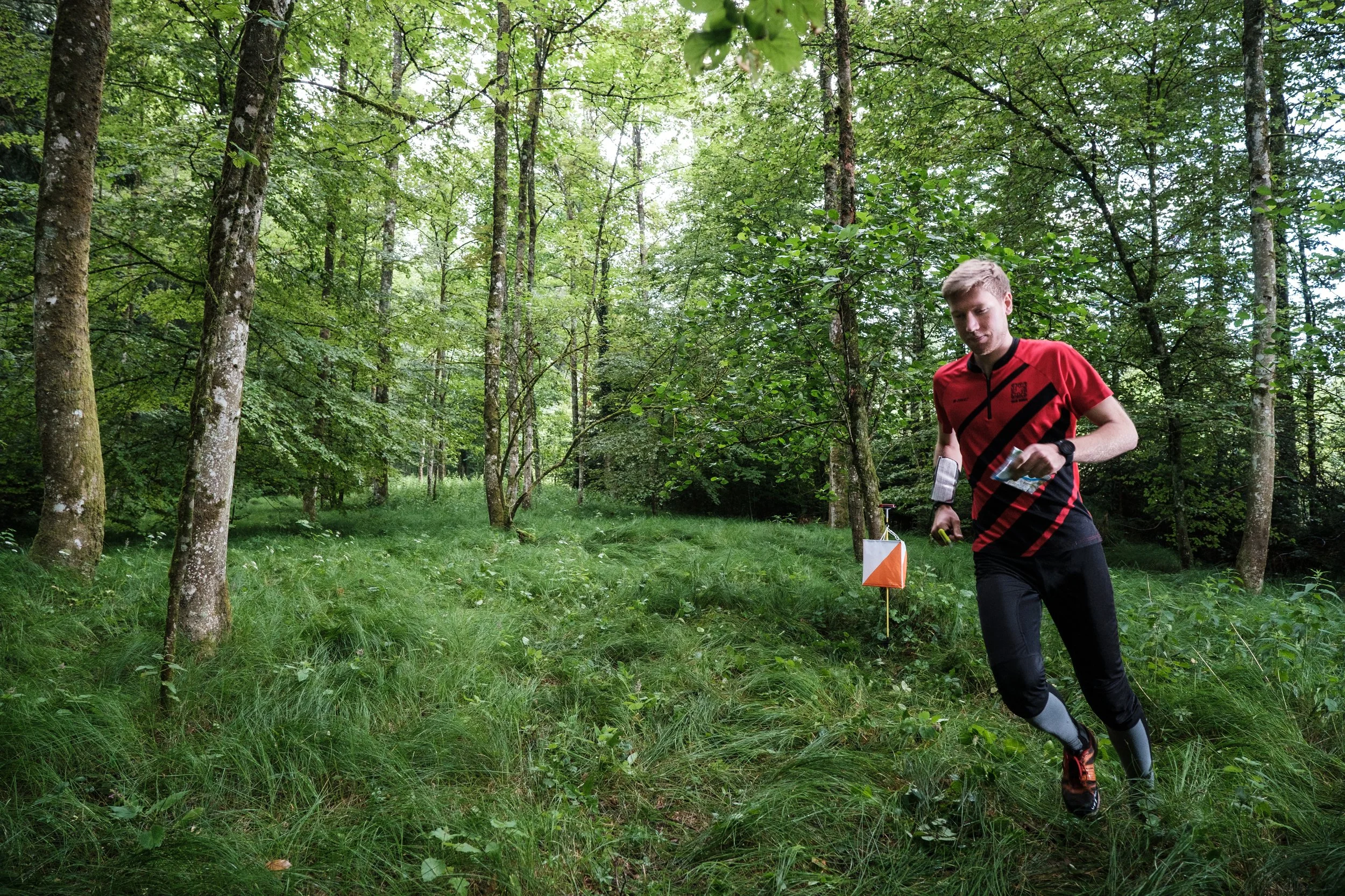 Ein Mann läuft im Wald, trägt eine rote und schwarze Sportkleidung und hält eine Karte in der Hand.