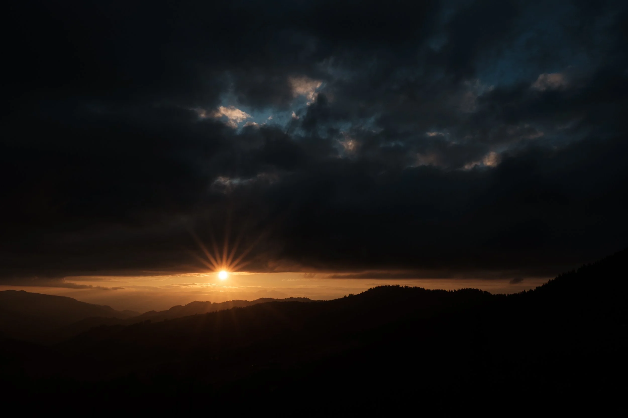 Sonnenuntergang hinter Bergen, dunkle Wolken am Himmel, Lichtstrahlen der Sonne scheinen durch Wolken. 