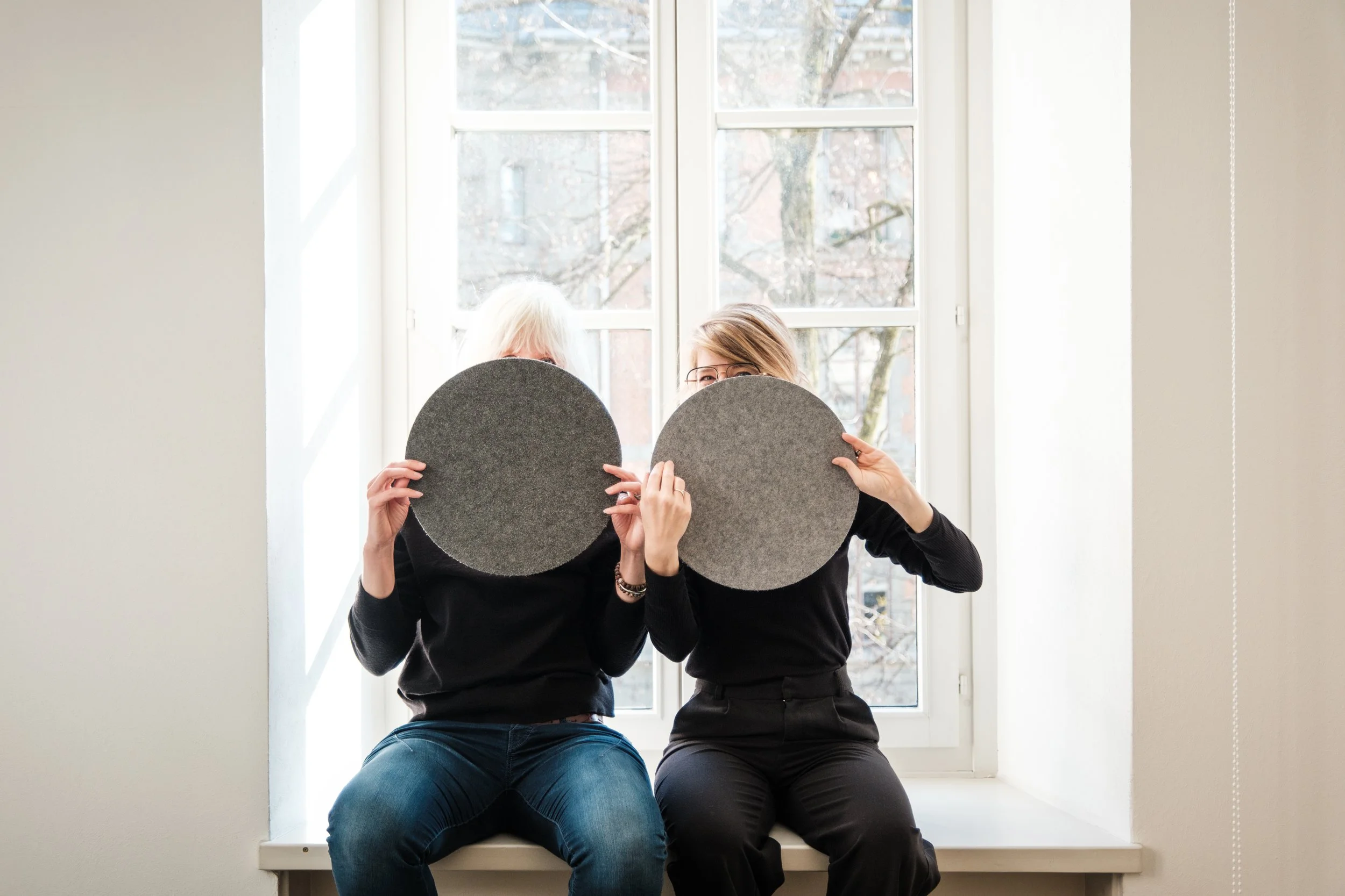 Zwei Frauen sitzen auf einer Fensterbank, vor einem großen Fenster mit Blick auf Bäume, und halten Filzunterlagen vor ihre Gesichter.