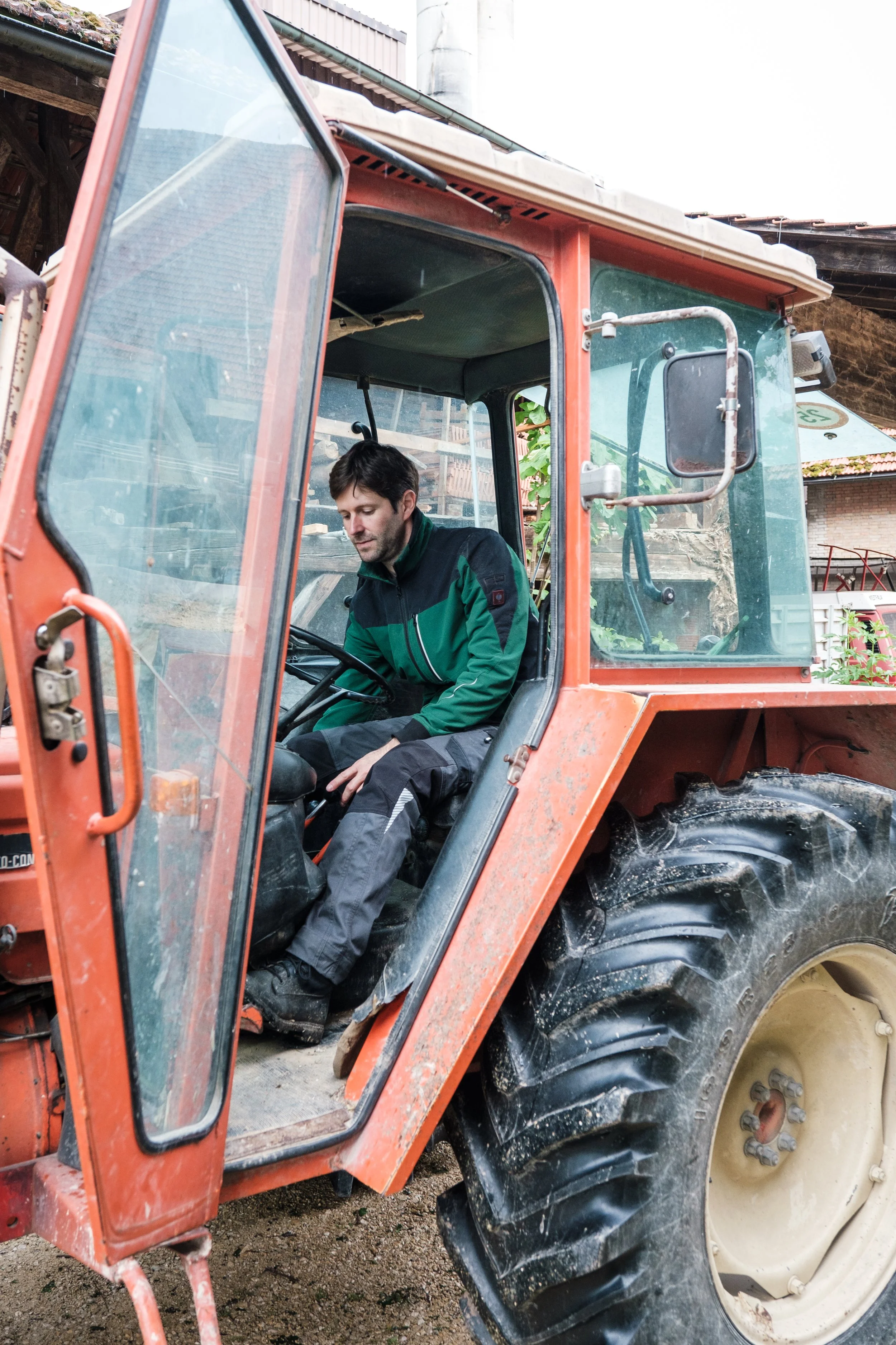 Mann sitzt im Führerhaus eines orangefarbenen Traktors, mit geschlossener Tür, im Freien auf einem Bauernhof oder Feld. Hintergrund mit Gebäuden und Pflanzen.