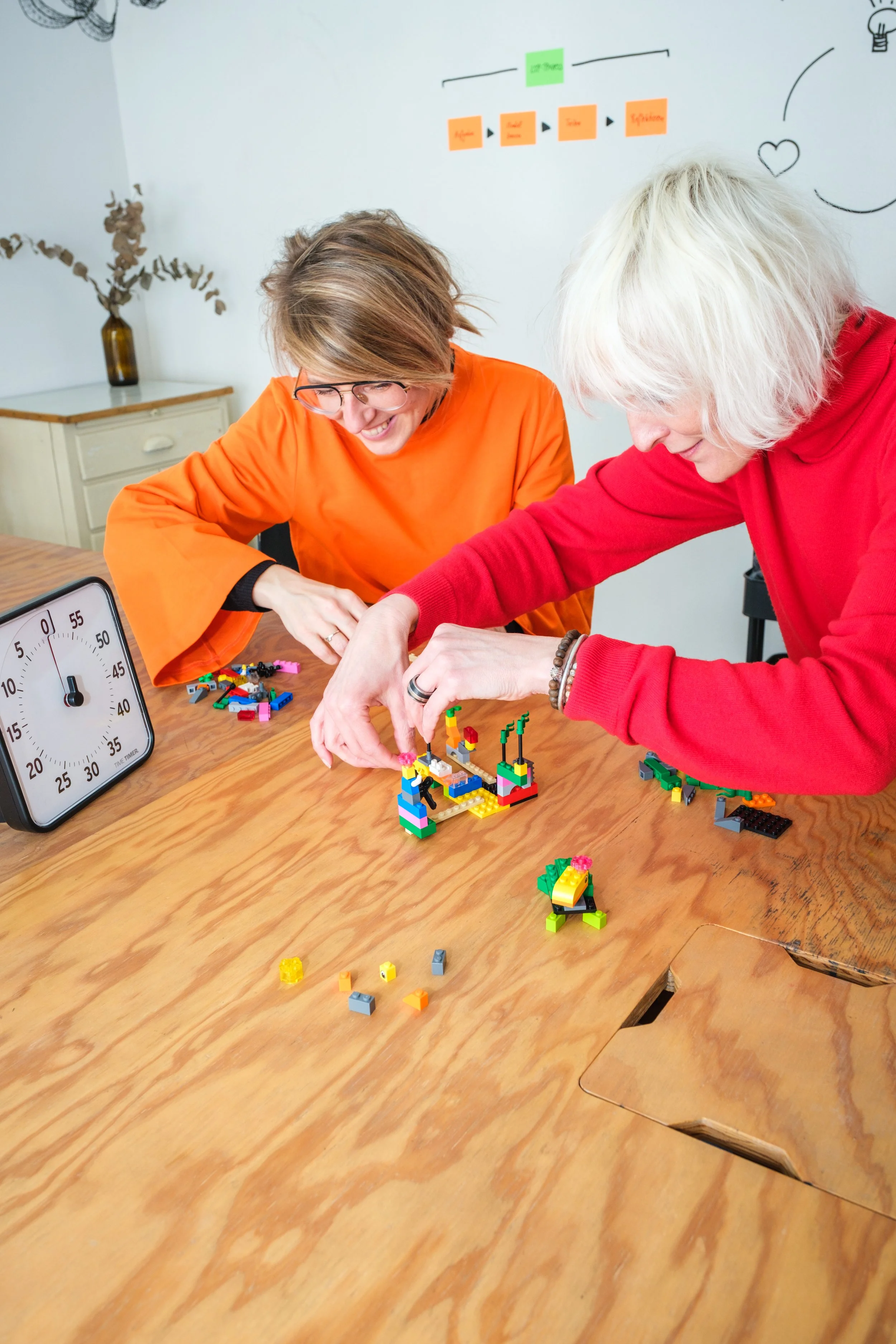 Zwei Frauen spielen zusammen mit Lego Steinen auf einem Holztisch, lachen und bauen ein LEGO-Modell. Auf dem Tisch steht eine Uhr. Im Hintergrund ist eine weiße Wand mit Post-its und Zeichnungen.