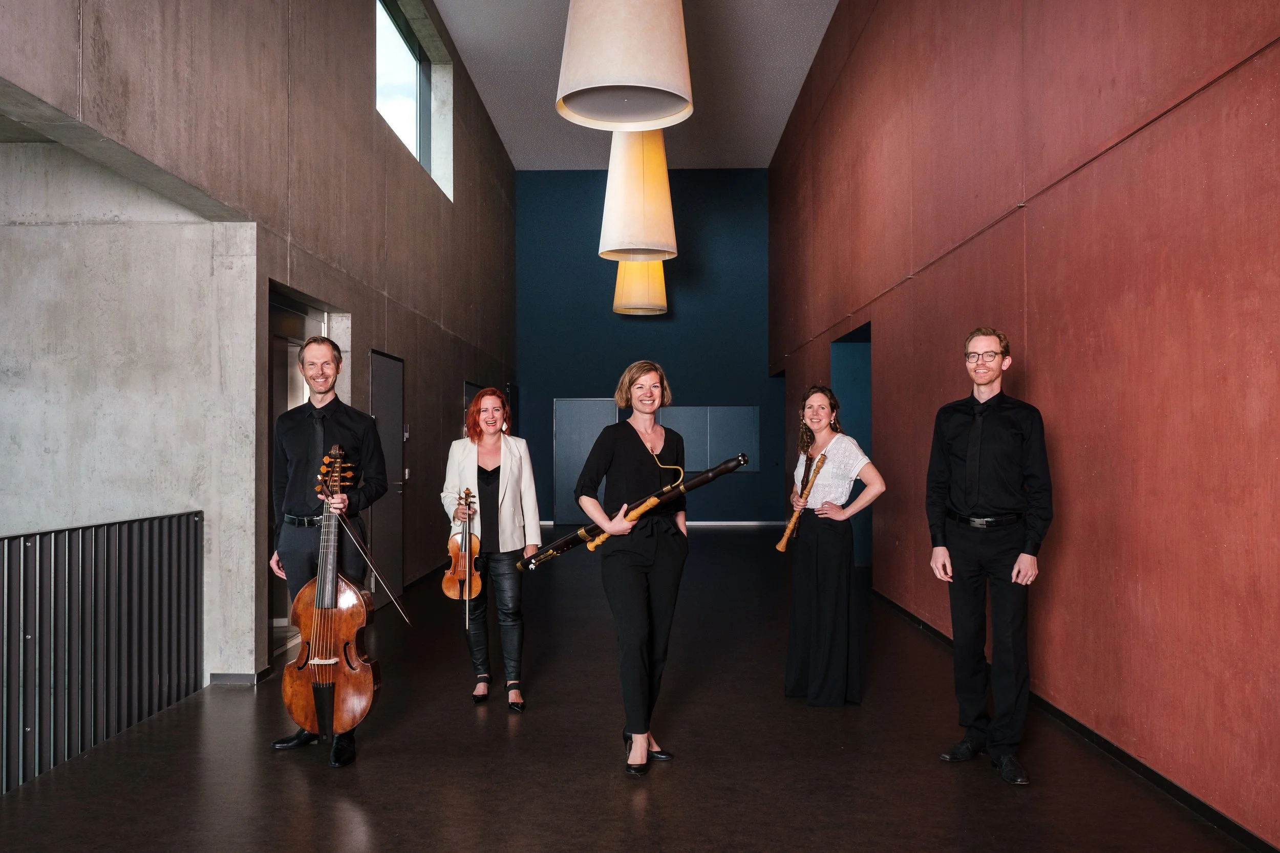 Fünf Musiker mit verschiedenen Instrumenten im modernen Flur, die lachen und posieren.
