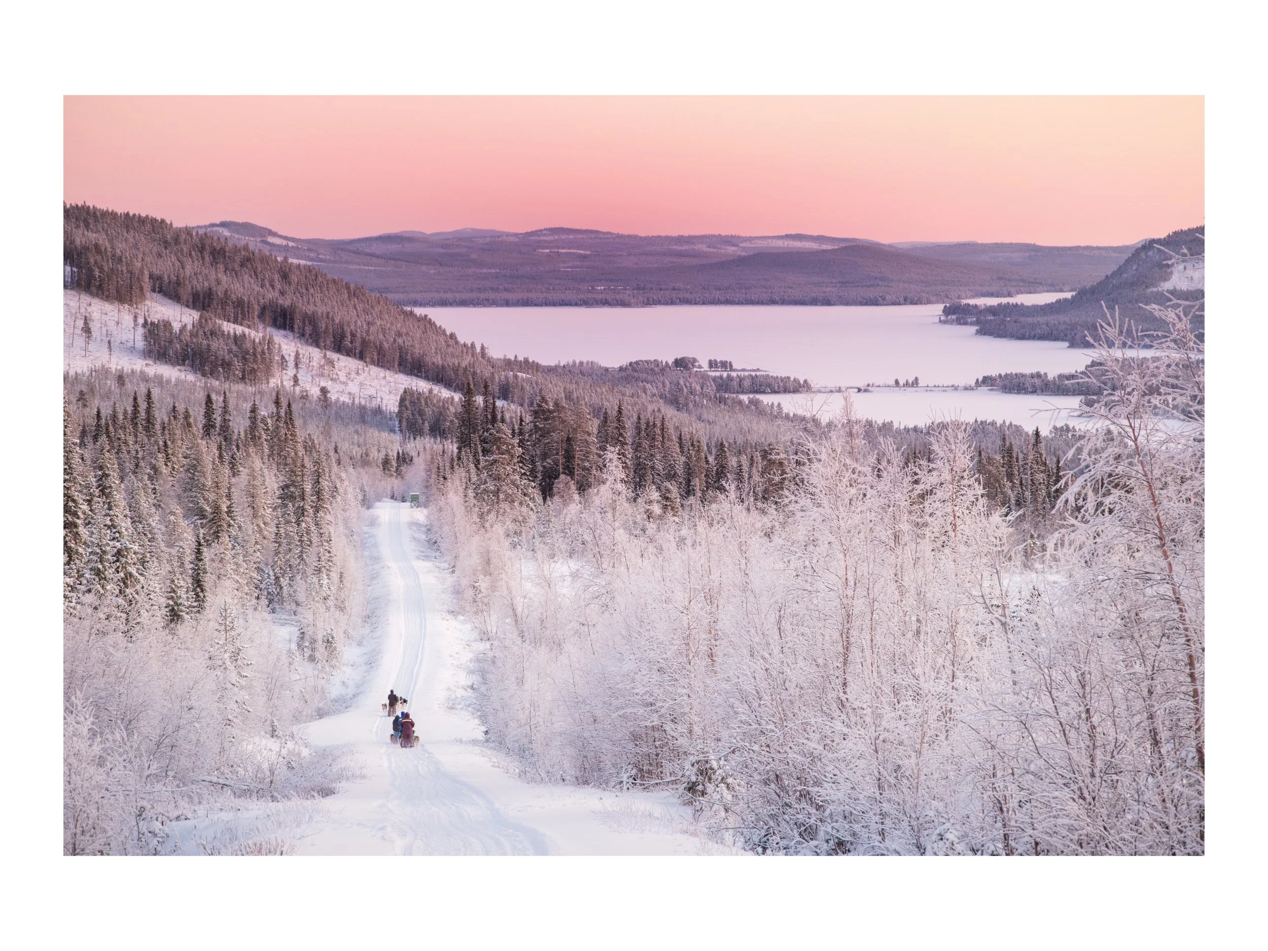 Schneebedeckte Winterlandschaft mit einem verschneiten Weg, auf dem Schlittenfahrer fahren, umgeben von gefrorenen Bäumen, mit einem See und Berge im Hintergrund bei Sonnenuntergang.