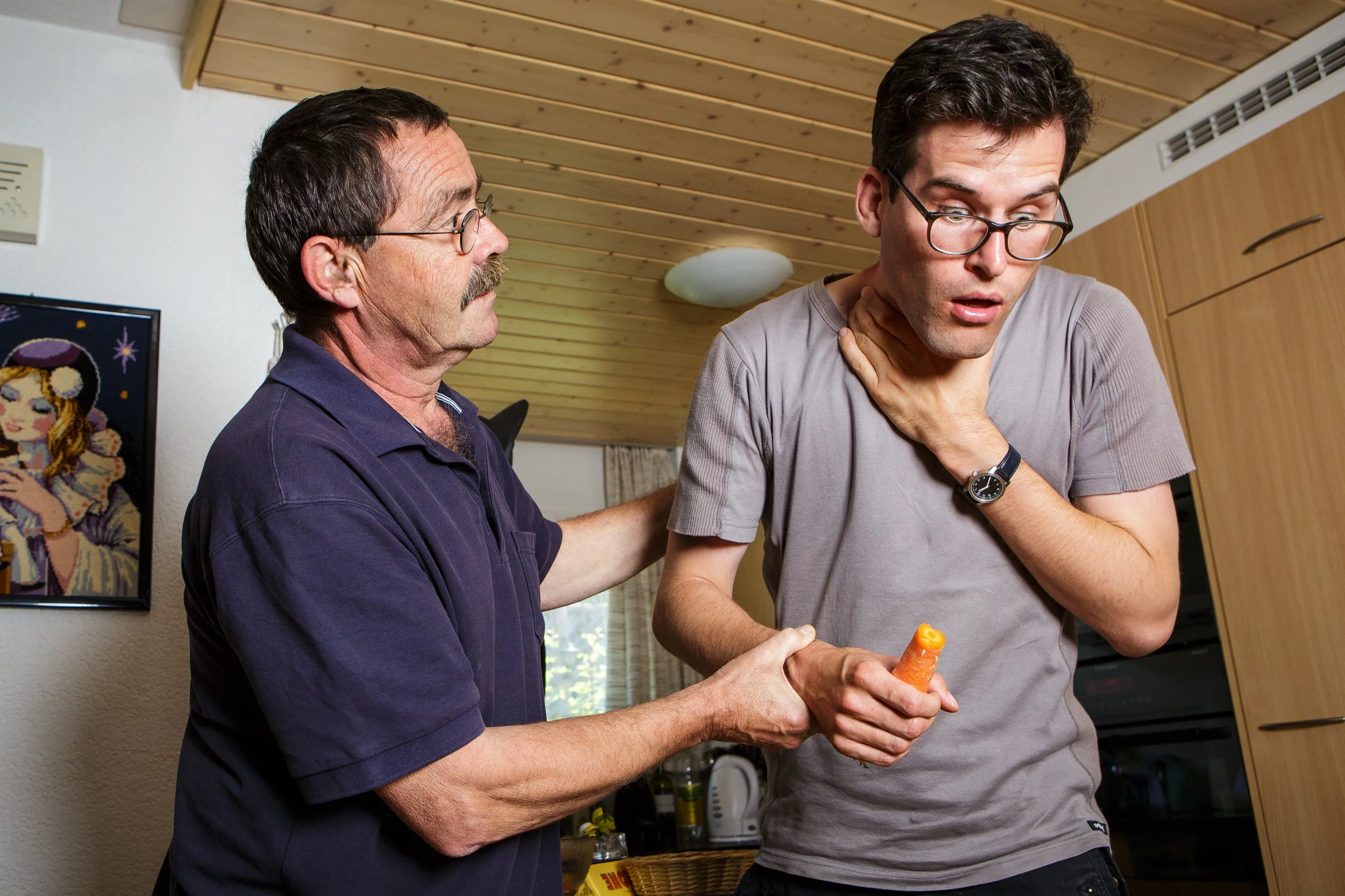 Ein älterer Mann hält einen jungen Mann am Hals, während der junge Mann mit einer Karotte in der Hand erschrocken aussieht.