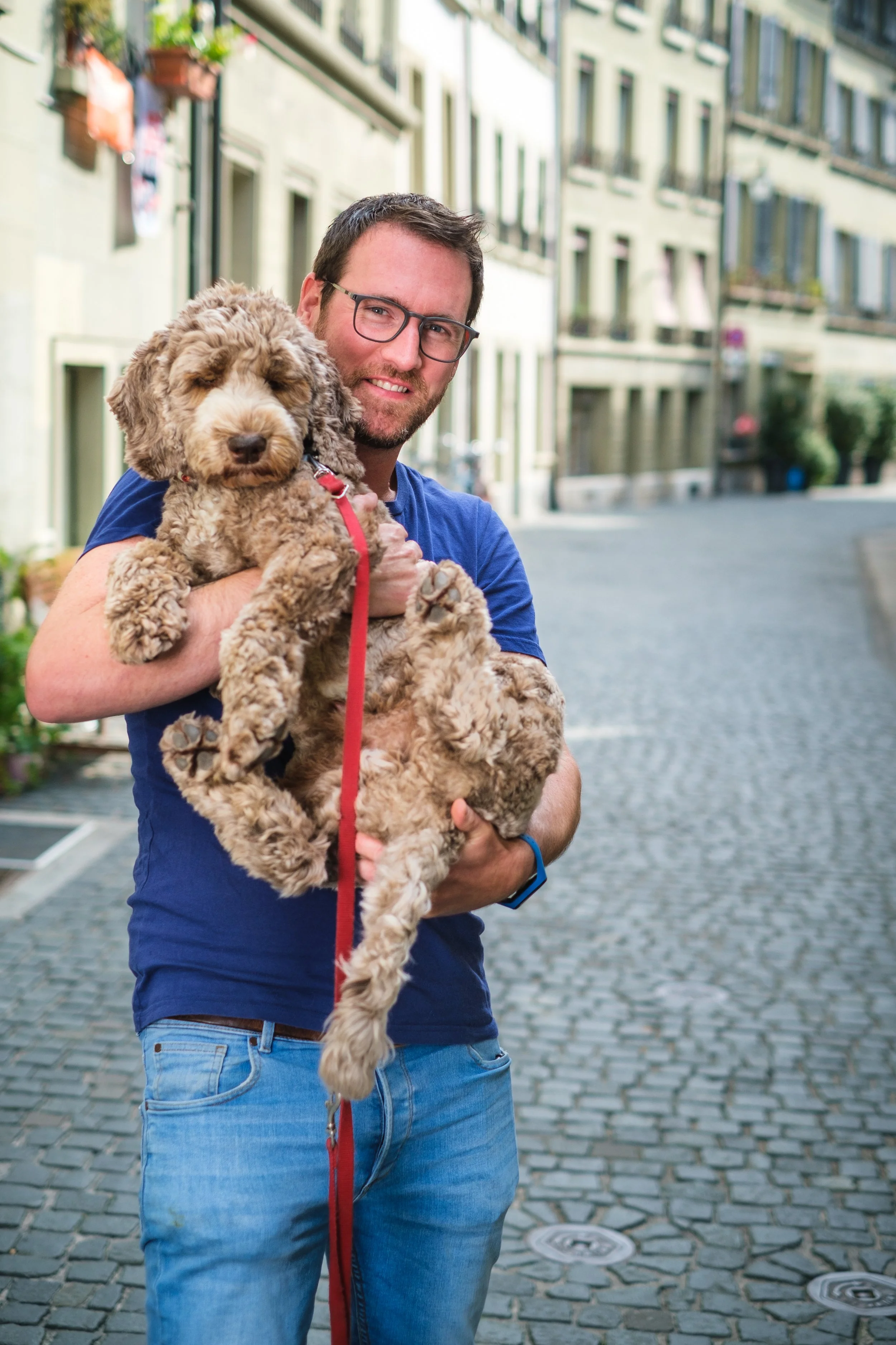 Ein Mann mit Brille hält ein braunes, lockiges Hundewelpen auf einer Kopfhöhe, in einer Stadtstraße mit alten Gebäuden im Hintergrund.